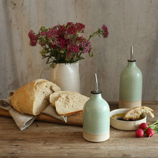 Small and large green oil pourers on table with slices of bread and a vase of wildflowers