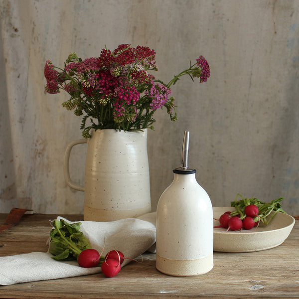 Small white oil pourer on table with bowl of radishes and jug of wildflowers