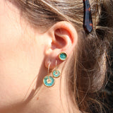 A women wearing a teal stud made from ceramic and glass with gold detailing, with two gold hoop earrings with matching round charms in small and large. 