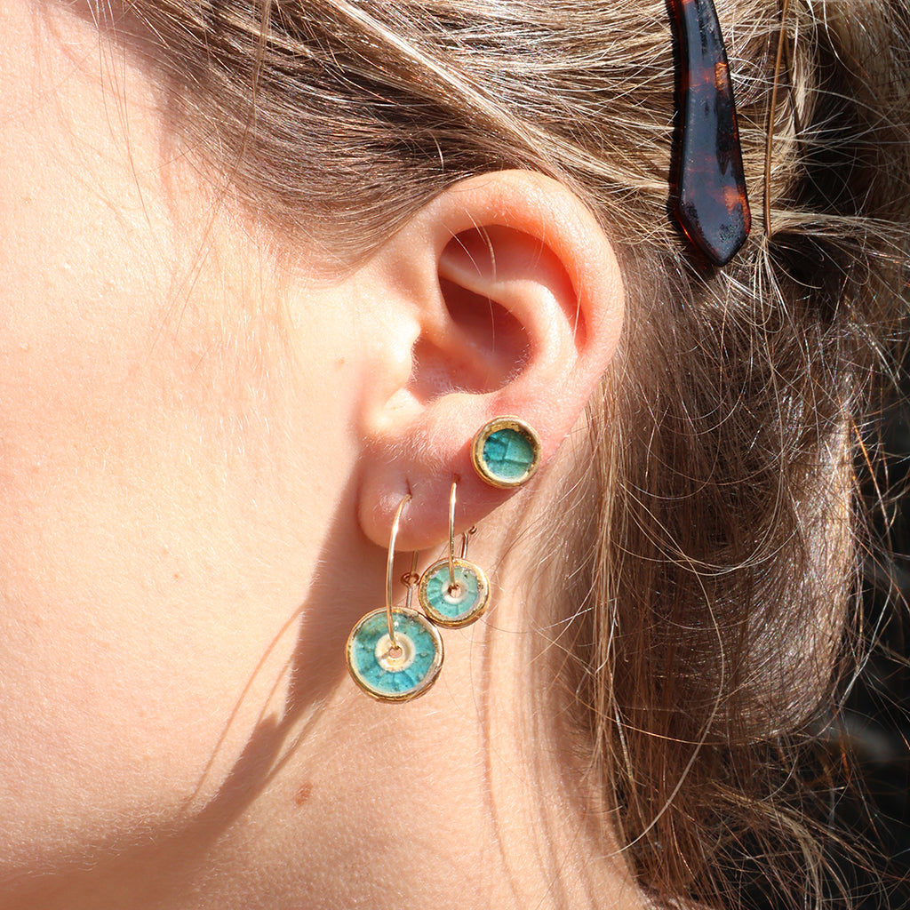 A women wearing a teal stud made from ceramic and glass with gold detailing, with two gold hoop earrings with matching round charms in small and large. 