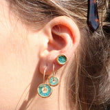A women's ear wearing gold fill hoop earrings with teal-coloured charms made from ceramic and glass, and a matching round stud earring. 
