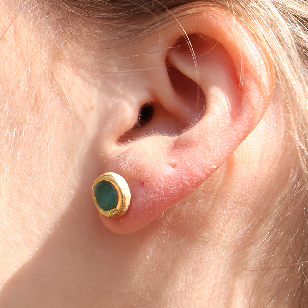 A close-up of a person's ear wearing a teal stud made from ceramic and glass with gold detailing. 