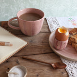 A pink stripe ceramic mug filled with coffee sits on a wooden table, alongside a soft-boiled egg in a matching egg cup, a plate of toast, a notebook and a spoon. 