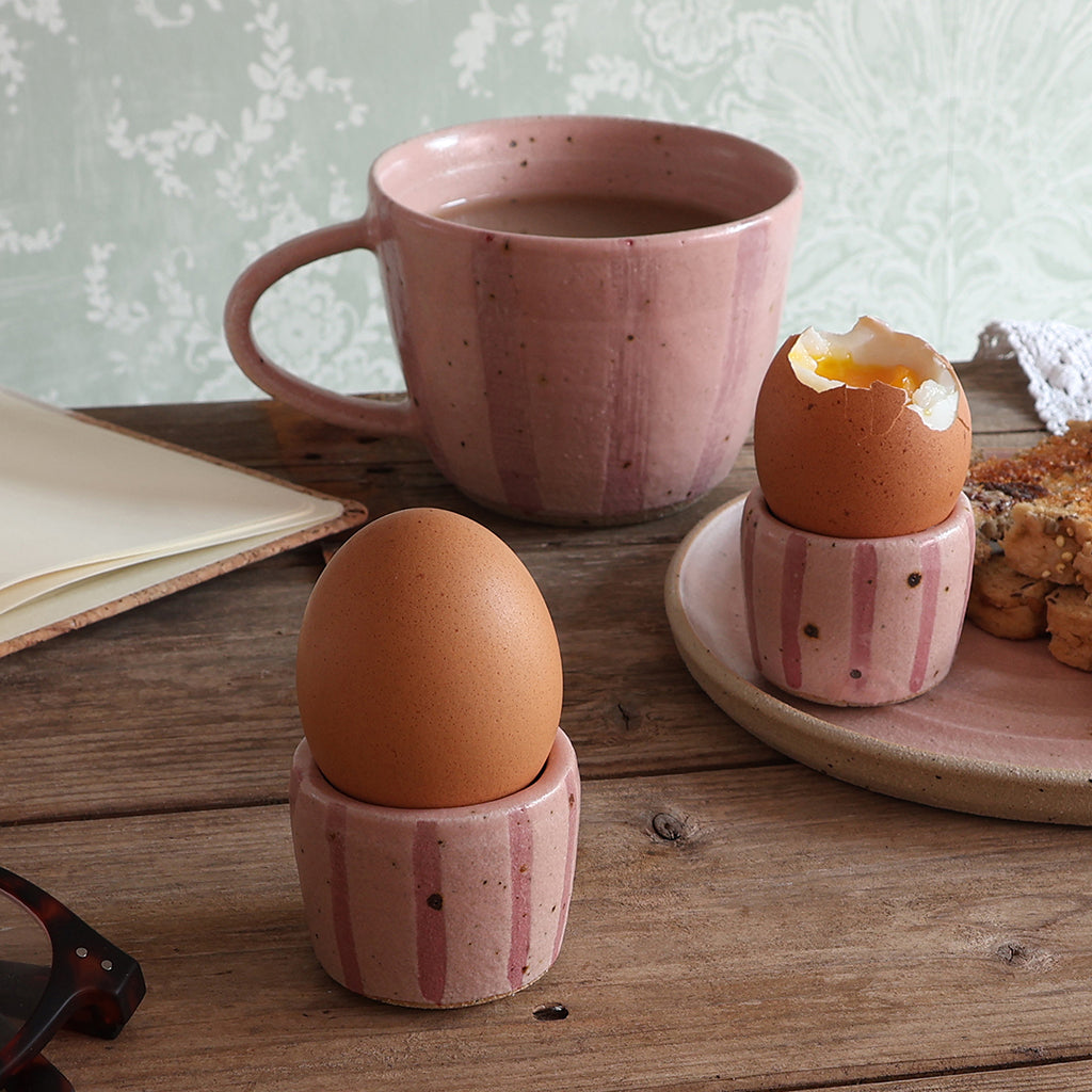 Two pink stripy ceramic egg cups holding boiled eggs sit on a rustic wooden table, with a matching pink mug and plate in the background.