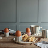 A styled breakfast scene on a wooden table featuring a blue and white stripe handmade ceramic mug, matching egg cups holding boiled eggs, toast, and a striped napkin against a muted blue paneled wall.