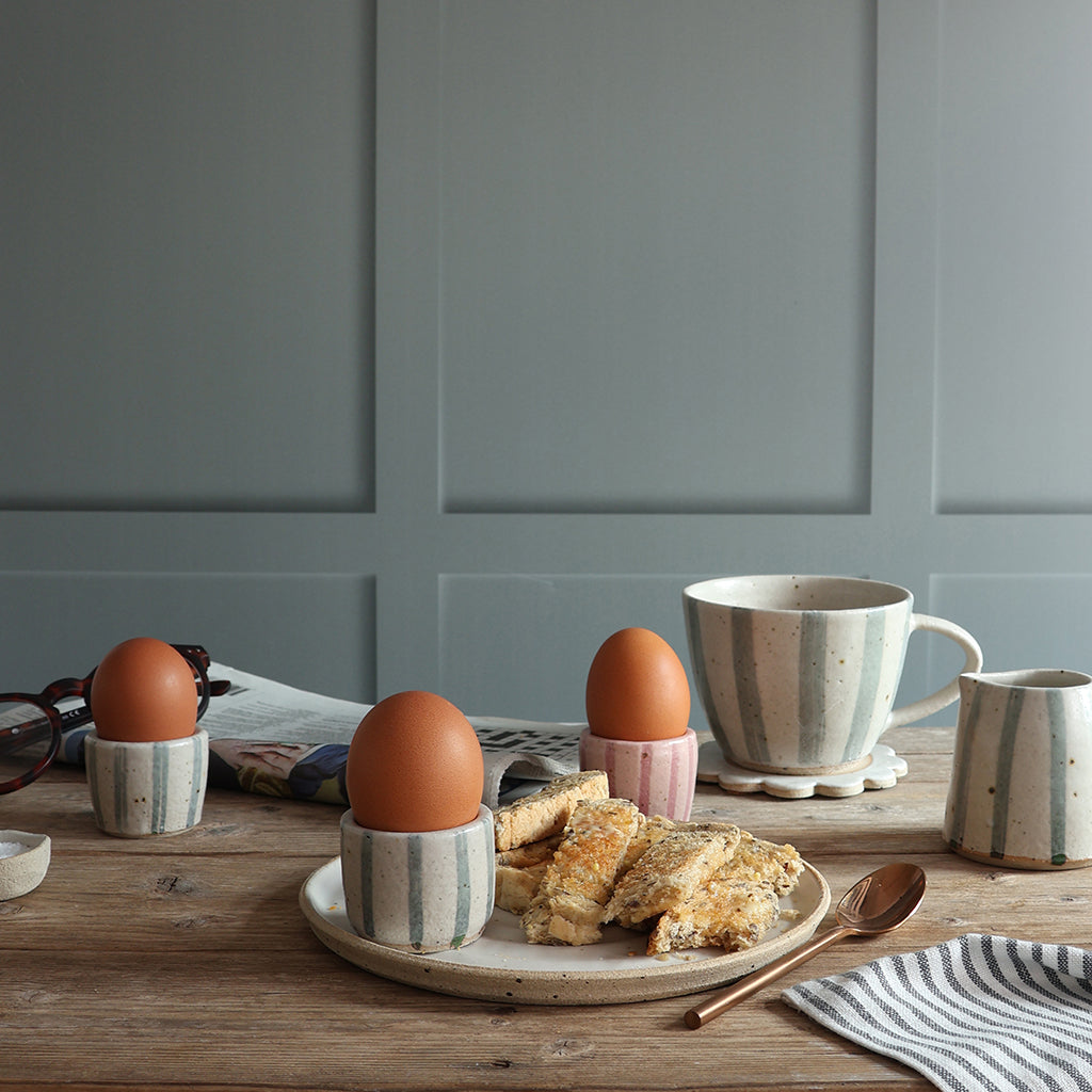 A styled breakfast scene on a wooden table featuring a blue and white stripe handmade ceramic mug, matching egg cups holding boiled eggs, toast, and a striped napkin against a muted blue paneled wall.