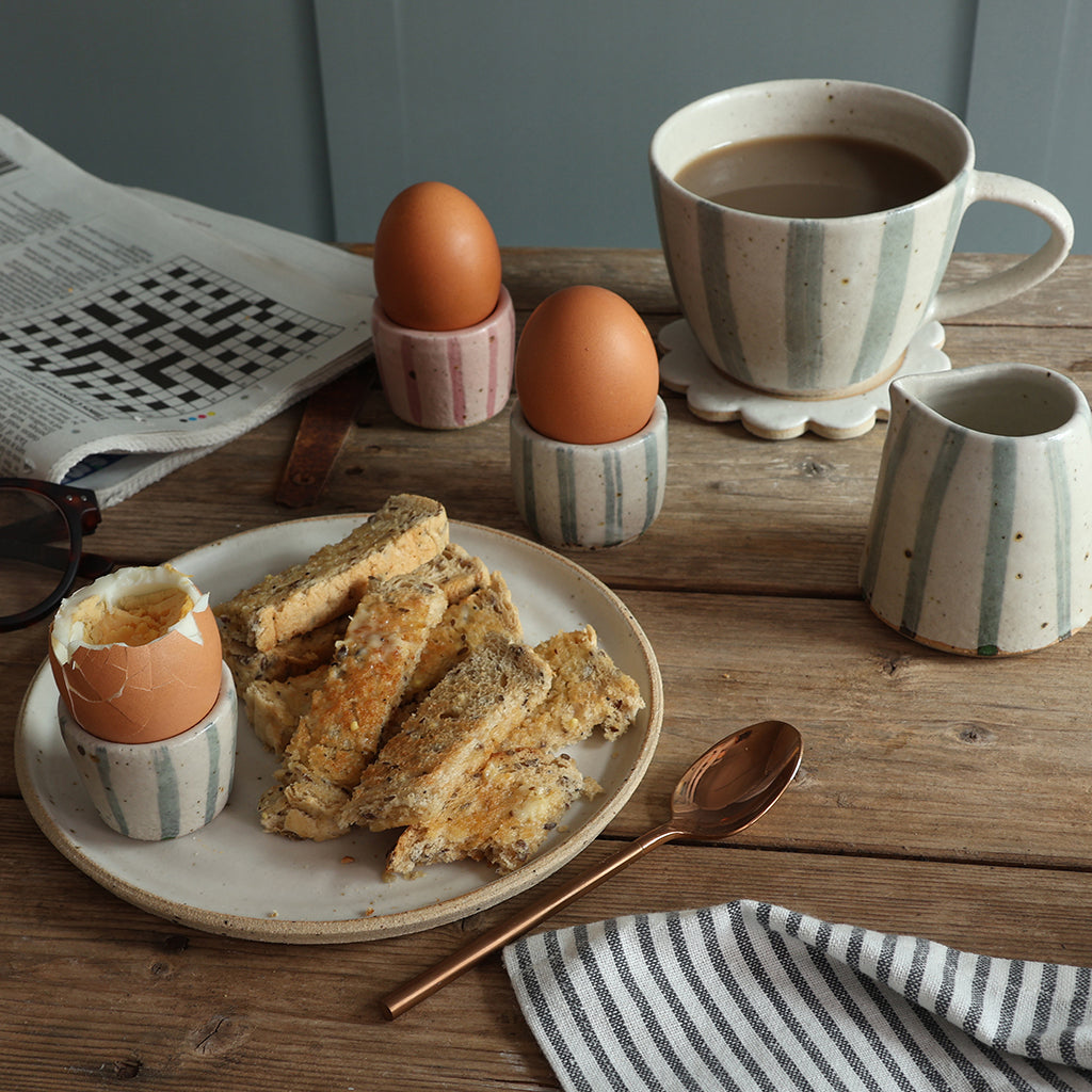 A styled breakfast scene on a wooden table featuring a blue and white stripe ceramic mug filled with coffee, a matching small jug, two boiled eggs in matching striped egg cups, a plate with toast, a spoon, newspaper, and a striped napkin. 