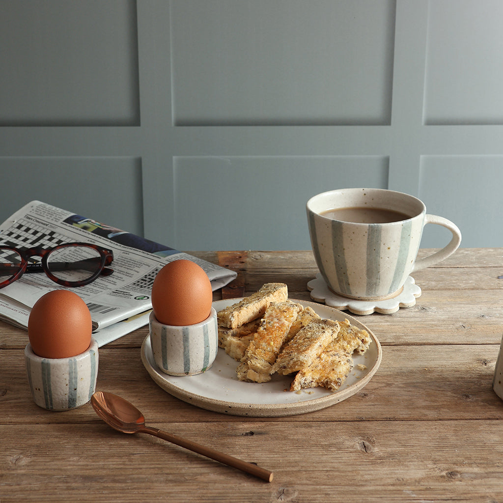 A styled breakfast scene on a wooden table with a handmade blue and white stripe ceramic mug of coffee on a coaster, a plate of toast, two eggs in matching striped egg cups, a spoon, and a folded newspaper against a paneled wall.
