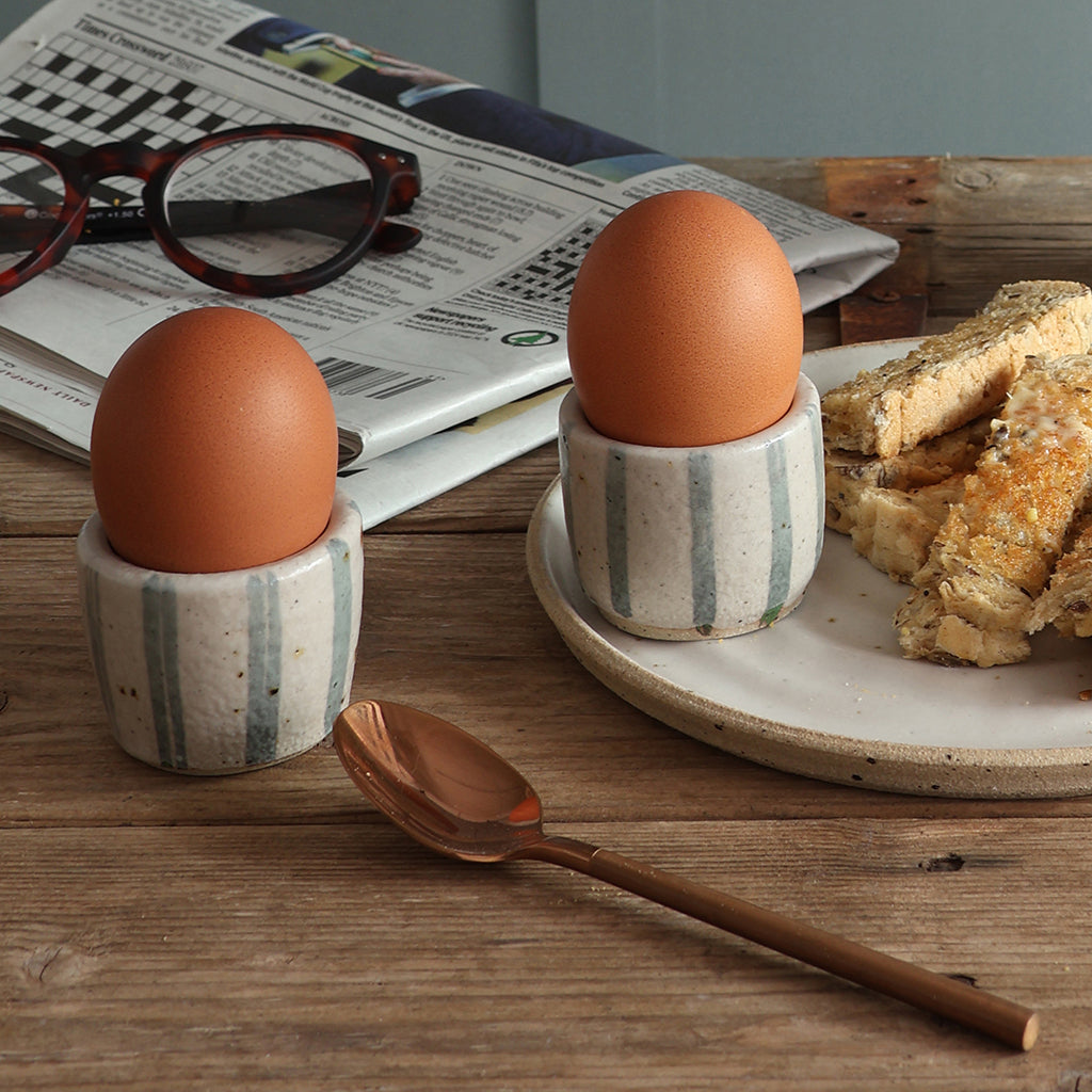 Two handmade blue and white stripe ceramic egg cups holding  boiled eggs on a rustic wooden table, with a plate of toast, a spoon, glasses, and a newspaper in the bacckground. 