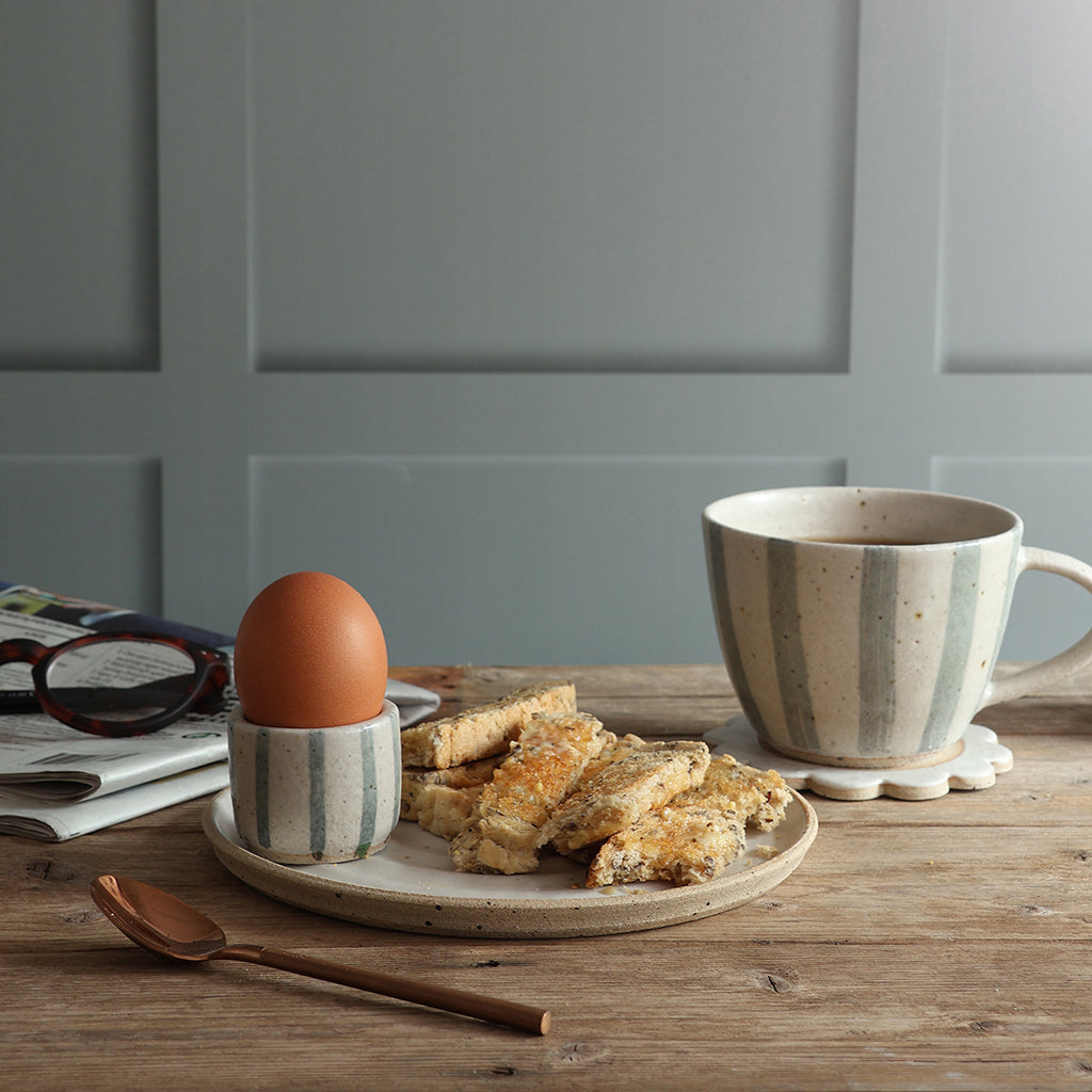 A handmade blue and white striped ceramic mug filled with coffee sits on a wooden table alonside a plate of toast, an egg in a matching egg cup, and a spoon, set against a paneled wall. 