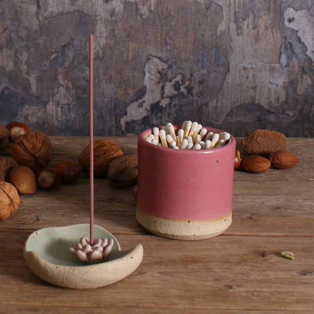 Ceramic incense holder with incense sticks and fuchsia match striker pot on a wooden surface with nuts in the background