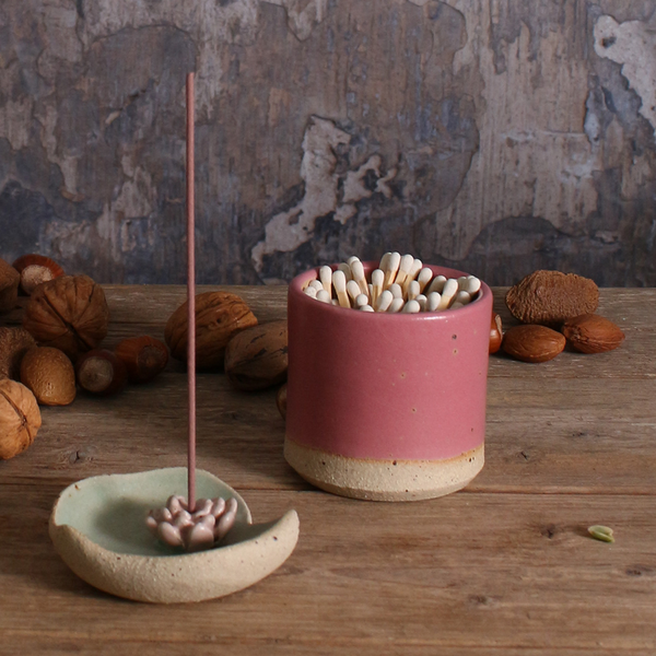 Ceramic incense holder with incense sticks and fuchsia match striker pot on a wooden surface with nuts in the background