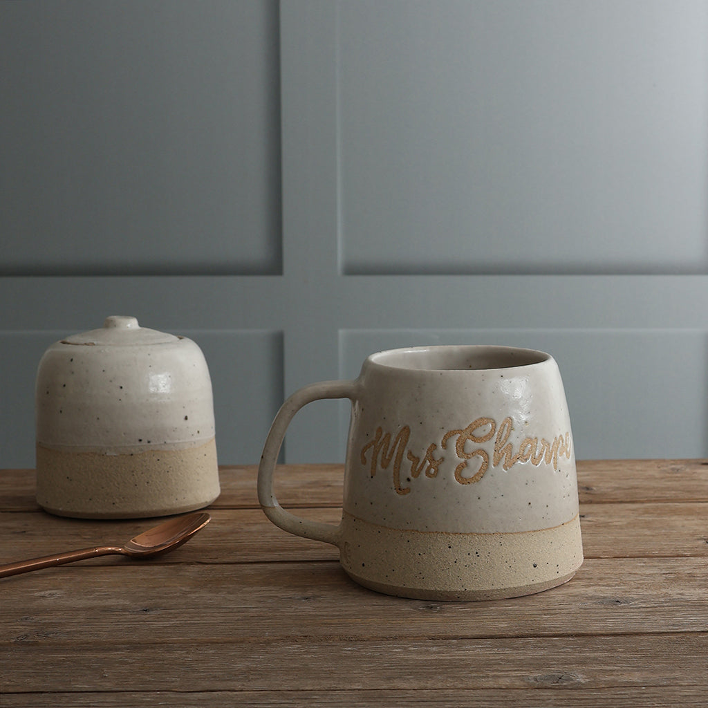 A handmade ceramic mug with a handle and a matching lidded sugar bowl sit on a rustic wooden table against a gray paneled wall. The mugis personalised with the name 'Mrs Sharp'. 