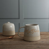 A handmade ceramic mug with a handle and a matching lidded sugar bowl sit on a rustic wooden table against a gray paneled wall. The mugis personalised with the name 'Mrs Sharp'. 