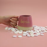 Pink mug with 'hot tea' text held by a hand on a pink background with scattered love heart sweets.