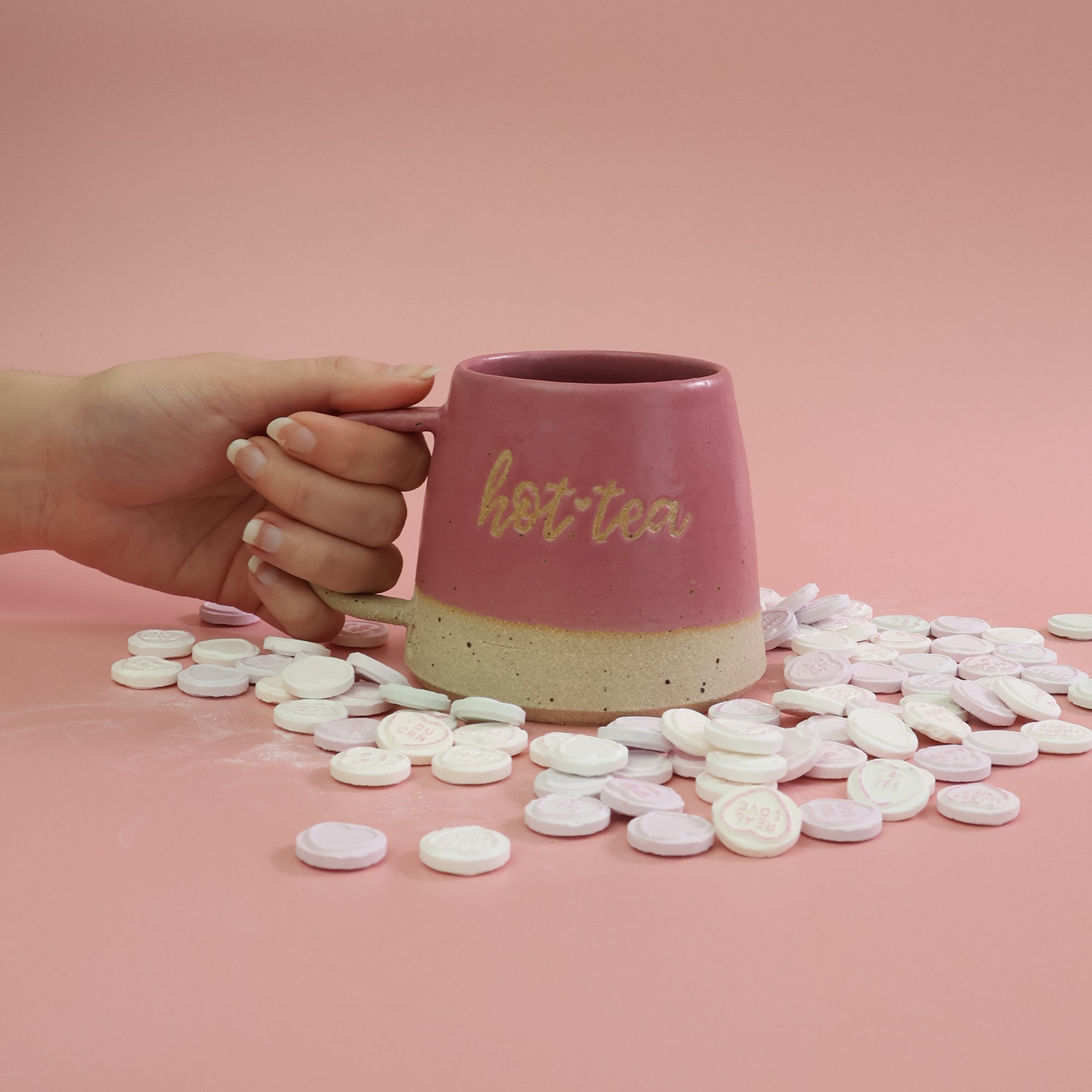Pink mug with 'hot tea' text held by a hand on a pink background with scattered love heart sweets.