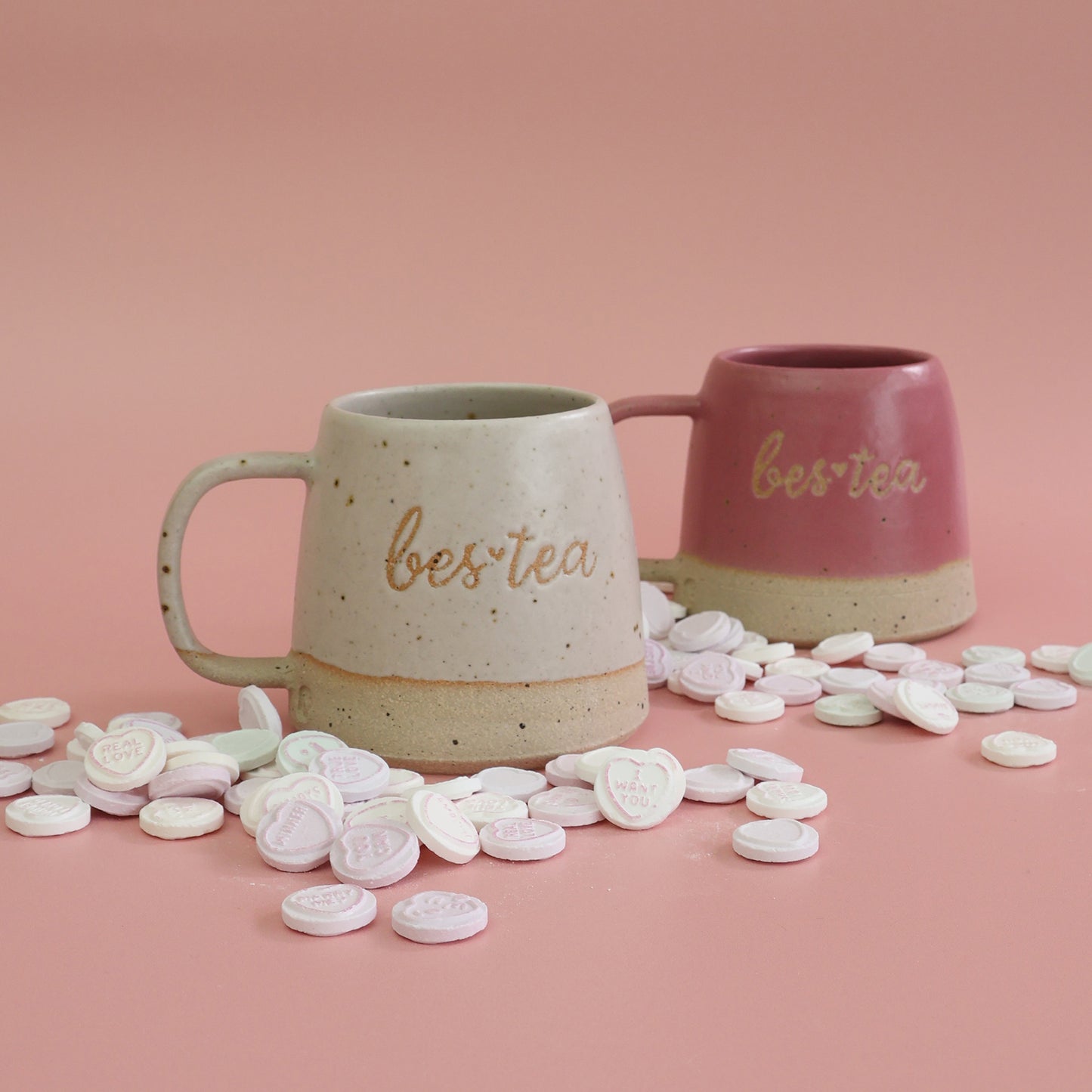 Two mugs with 'best tea' text on a pink background with heart-shaped candies.