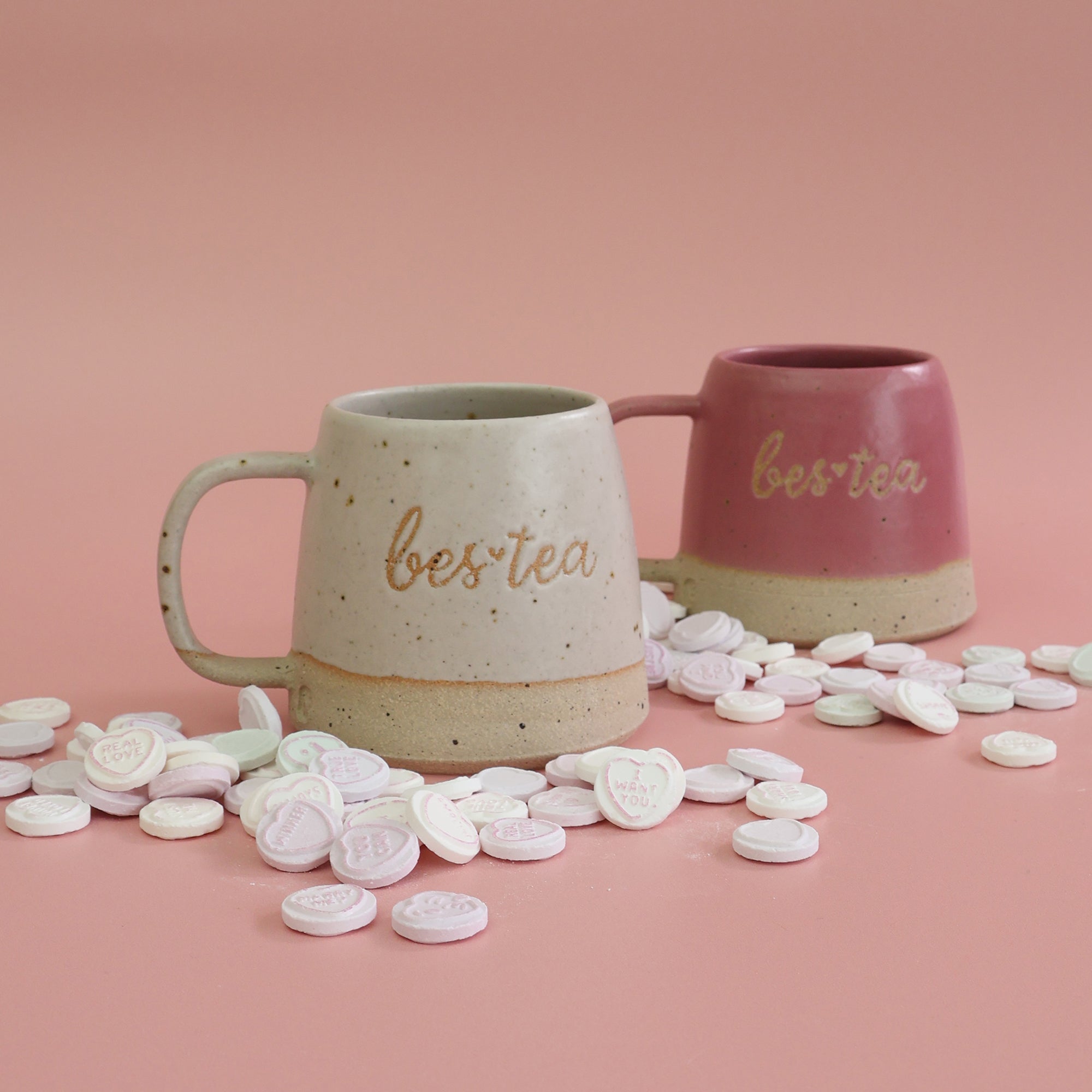 Two mugs with 'best tea' text on a pink background with heart-shaped candies.