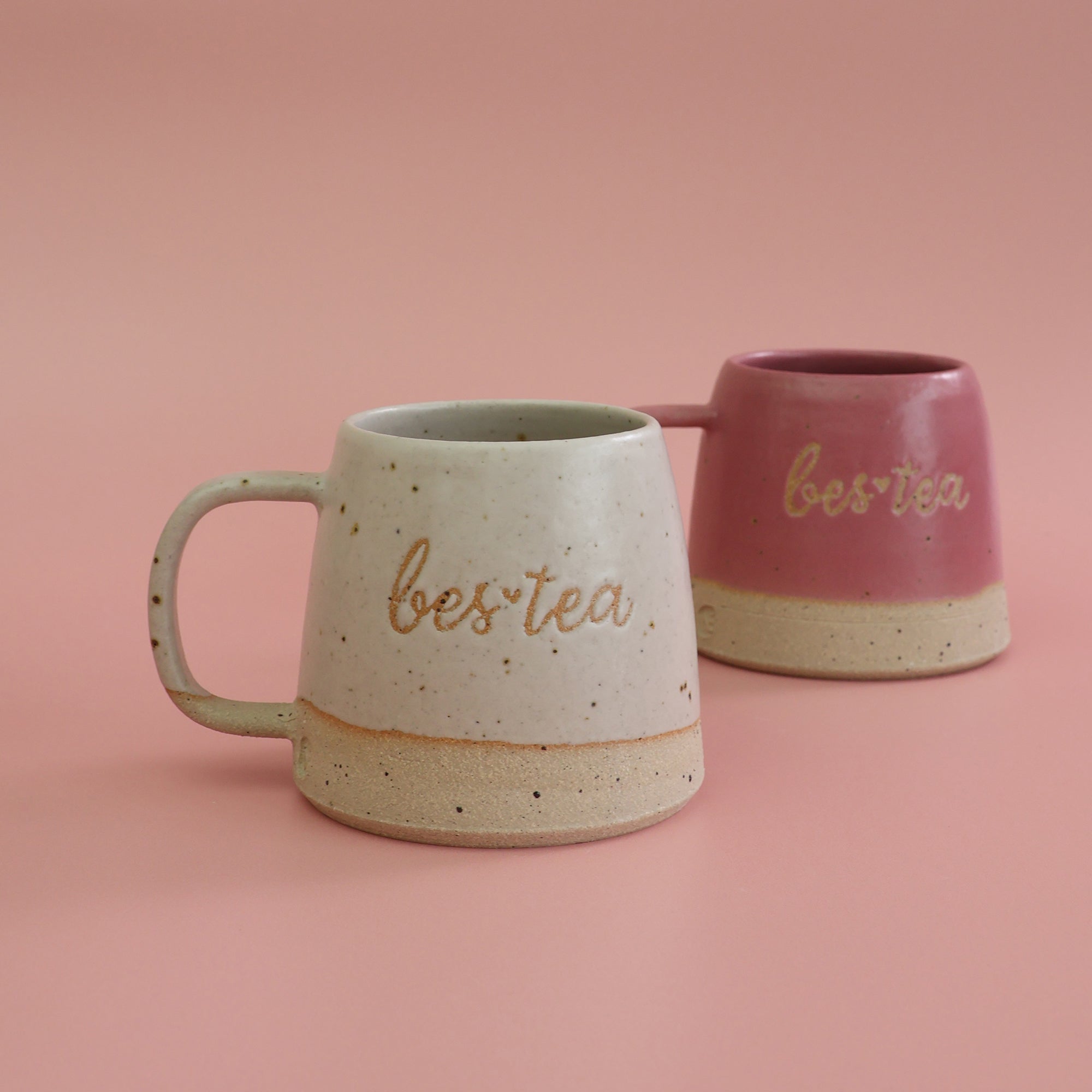 Two handmade ceramic mugs in bright pink and white with 'bes*tea' text on a pink background