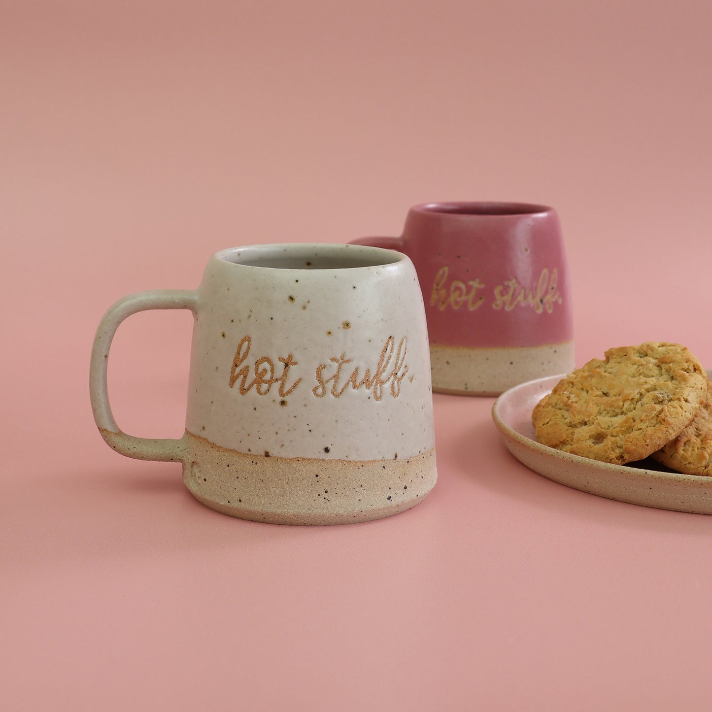 Two handmade ceramic mugs with 'hot stuff' text and a plate of cookies on a pink background. The mugs are in bright pink and white with clay flecks breaking through the glaze - mugs are handmade in the UK by Habulous Ceramics 