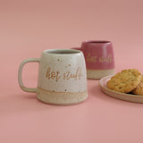 Two handmade ceramic mugs with 'hot stuff' text and a plate of cookies on a pink background. The mugs are in bright pink and white with clay flecks breaking through the glaze - mugs are handmade in the UK by Habulous Ceramics 