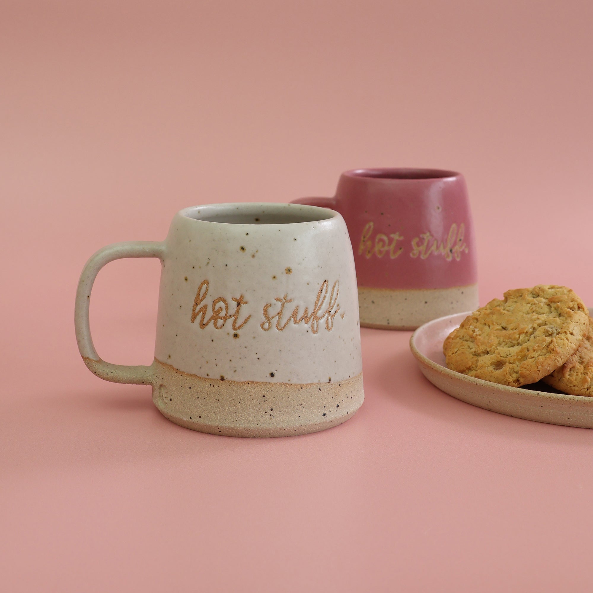 Two handmade ceramic mugs with 'hot stuff' text and a plate of cookies on a pink background. The mugs are in bright pink and white with clay flecks breaking through the glaze - mugs are handmade in the UK by Habulous Ceramics 