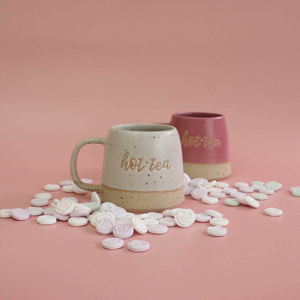 Two ceramic handmade hottie mugs featuring text 'hot tea' text on a pink background with heart-shaped love heart sweets

