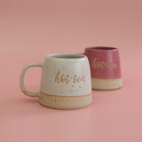 Two handmade stoneware ceramic mugs, one bright pink and the other white with 'hot tea' text a pun on the saying Hottie with a pink background