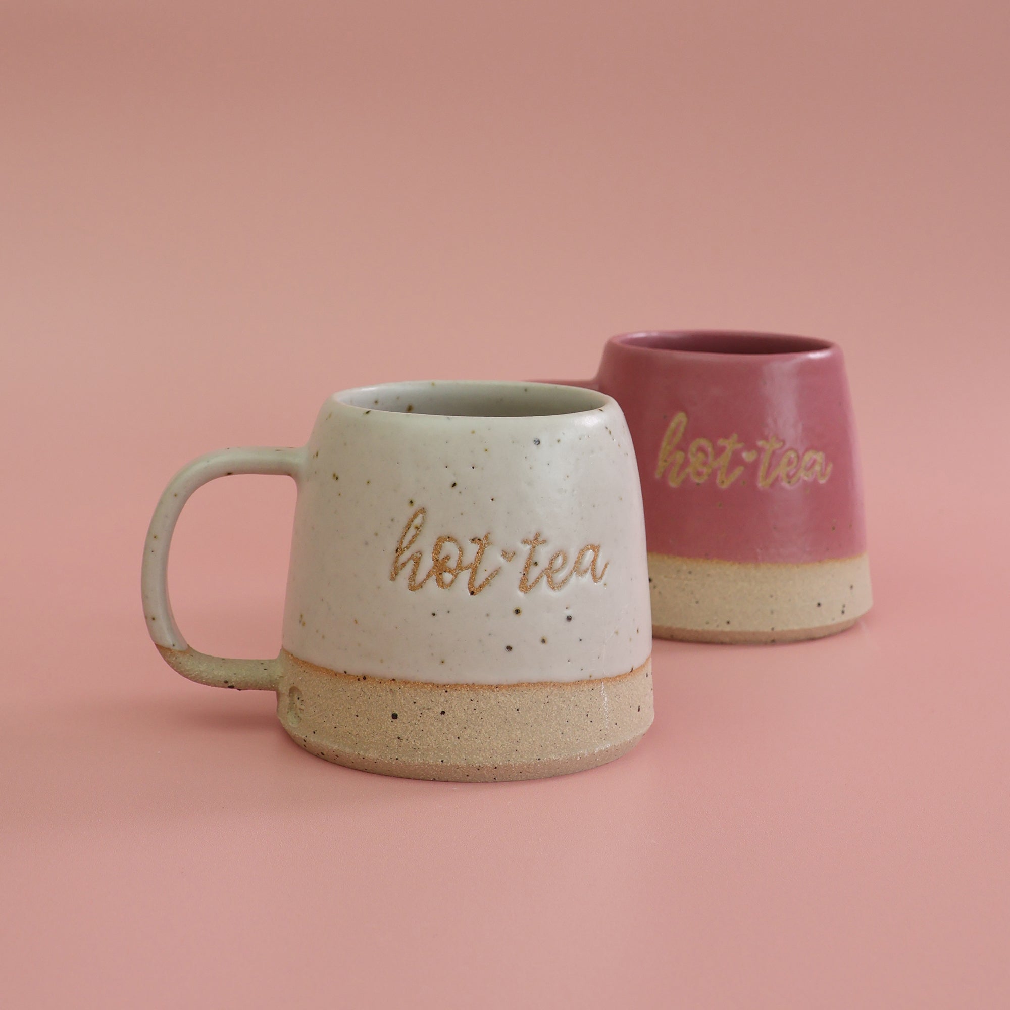 Two handmade stoneware ceramic mugs, one bright pink and the other white with 'hot tea' text a pun on the saying Hottie with a pink background
