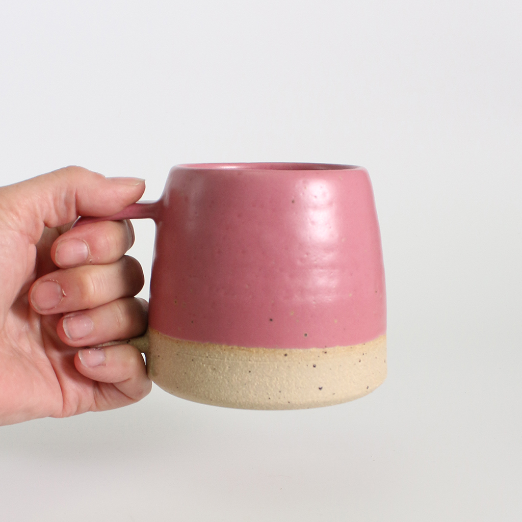 Hand holding a fuchsia pink ceramic mug with a beige base on a light grey background