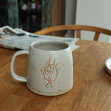 Ceramic mug with a handdrawn children's design on a wooden table with a newspaper in the background