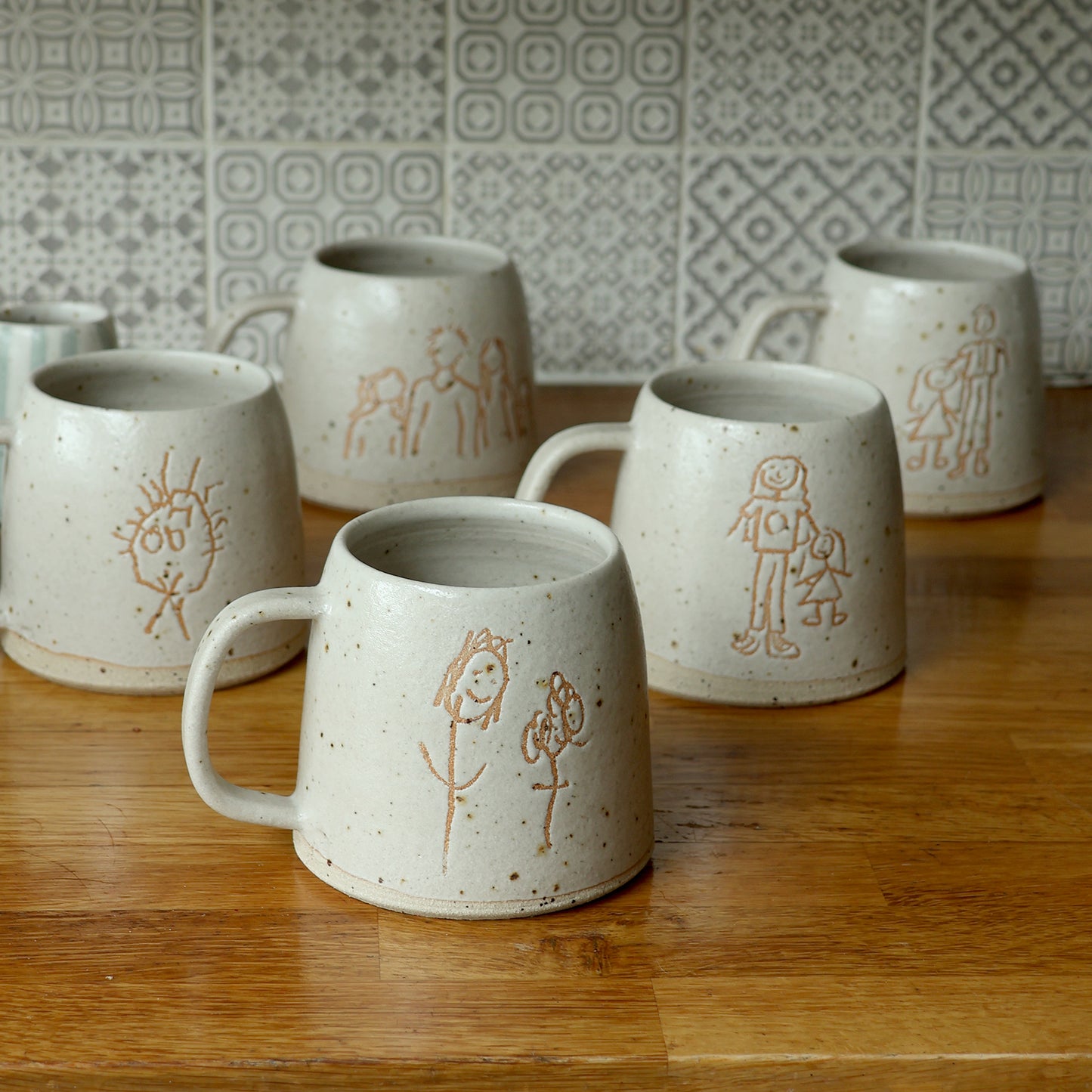 5 custom children's drawings ceramic mugs sat on a wooden surface featuring a range of drawings glazed in our off-white glaze.