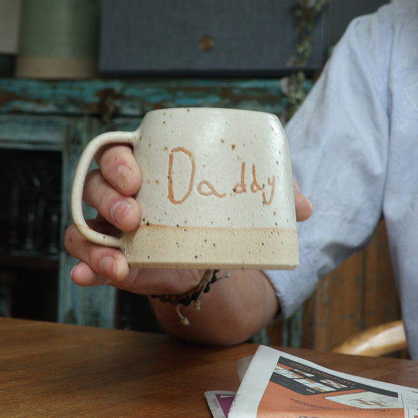 Person holding a ceramic mug with 'Daddy' engraving on a wooden table.