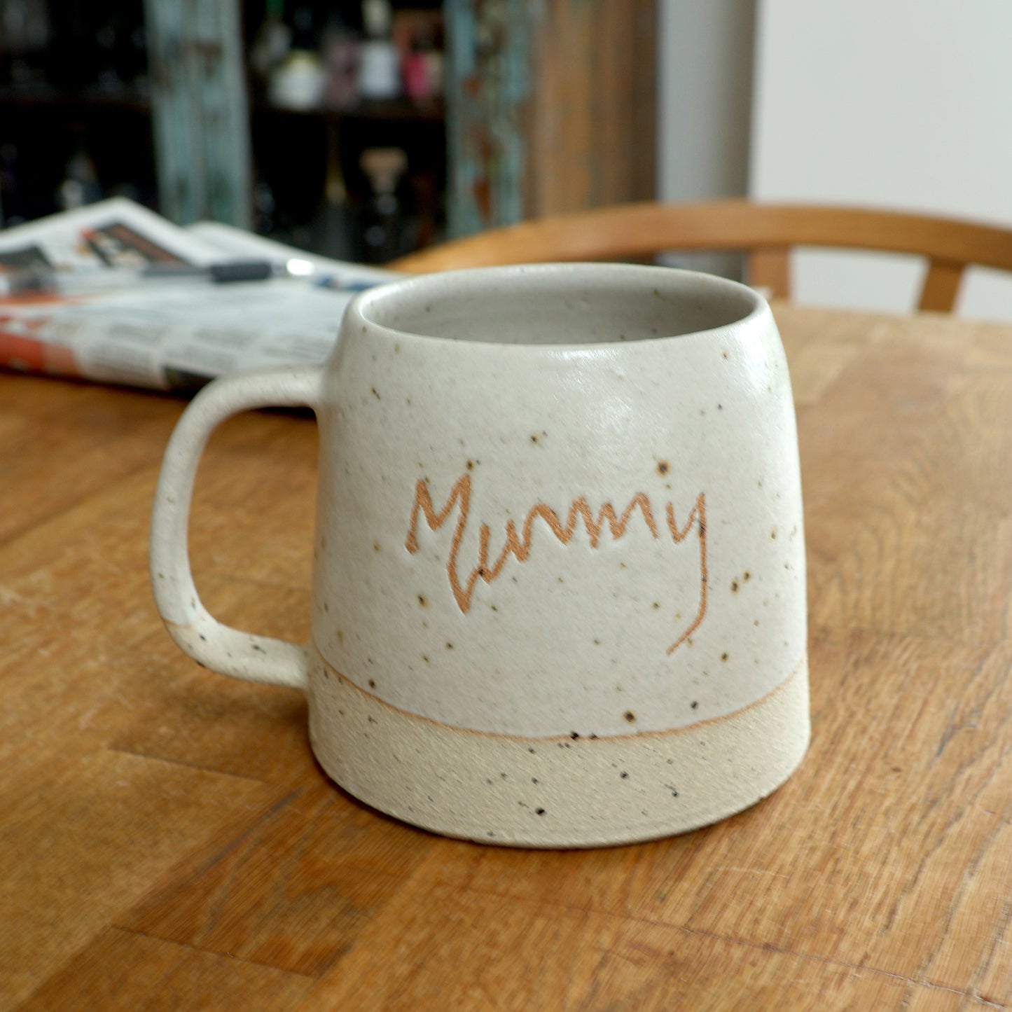 Handmade handwriting ceramic mug with 'Mummy' on a wooden table with a newspaper in the background