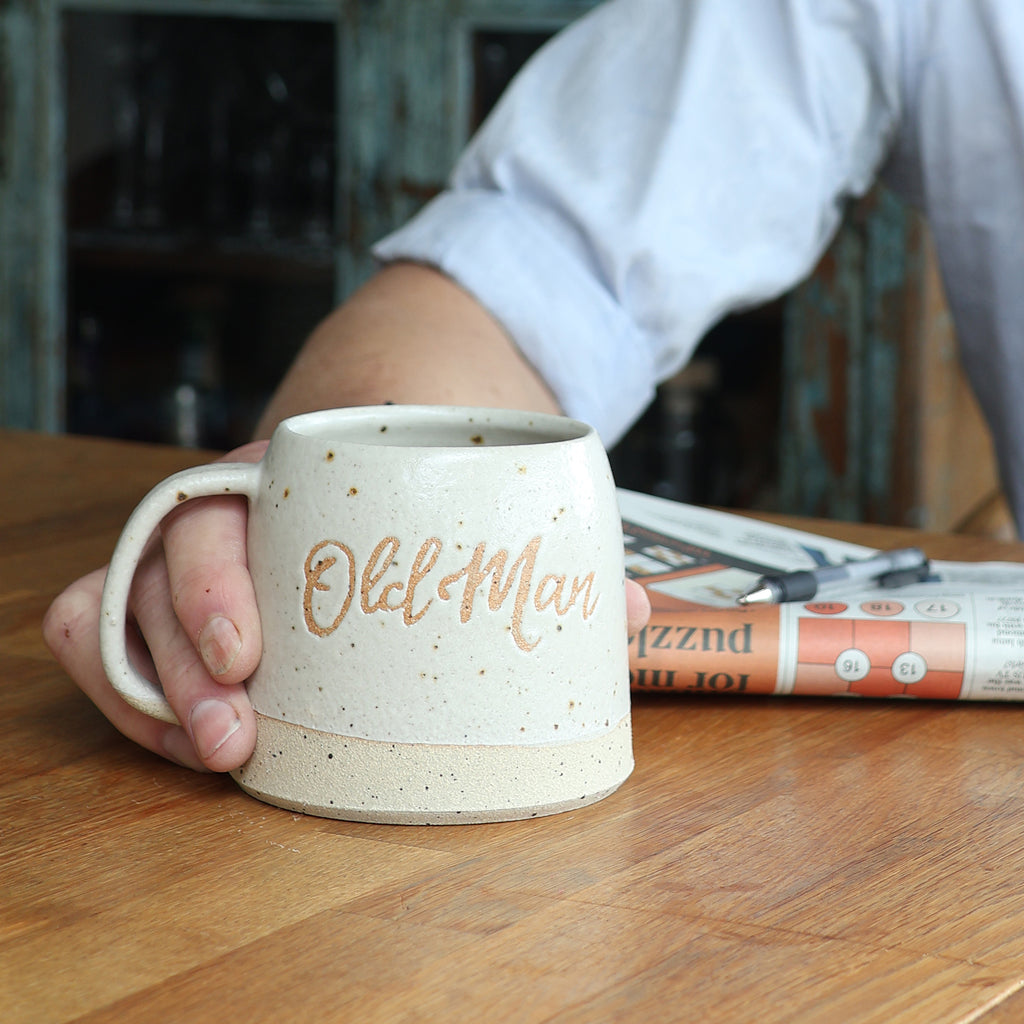 Person holding a ceramic mug with 'Old Man' text on a wooden table.