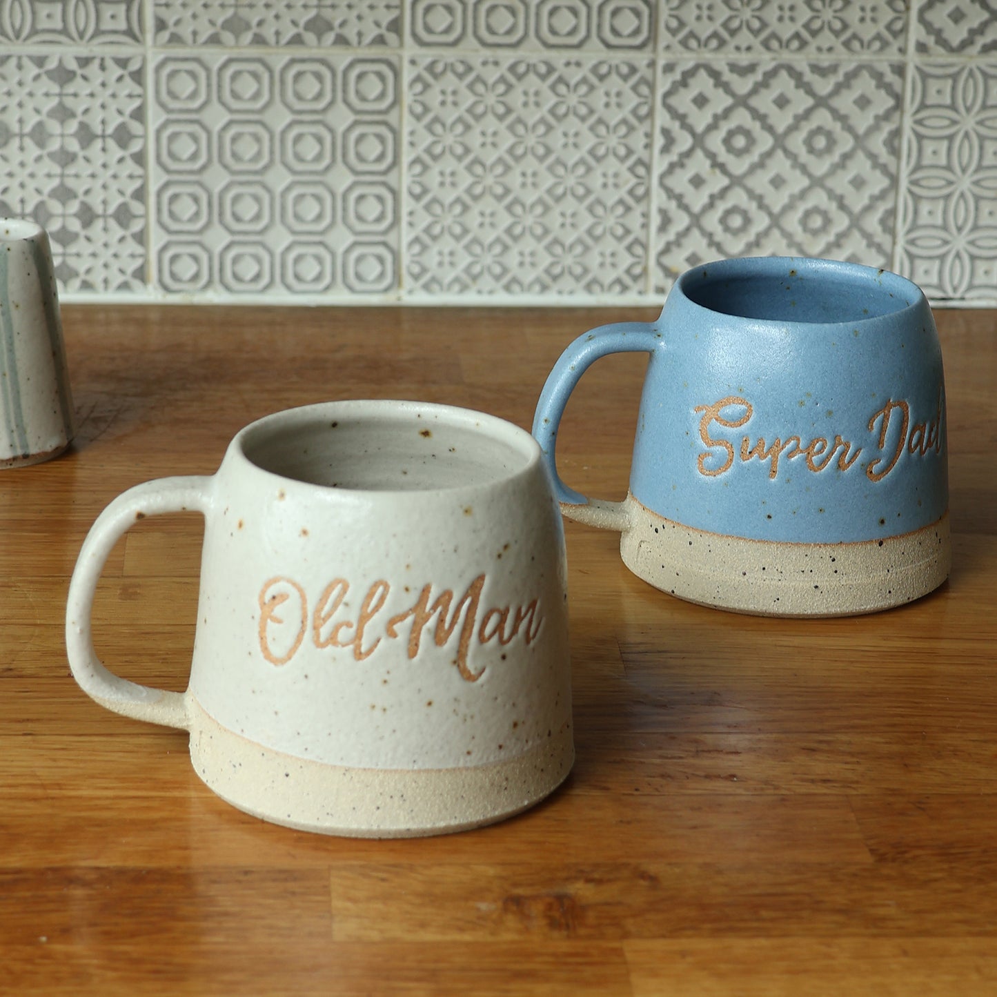 Two ceramic mugs with text on a wooden surface and tiled wall background