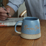 Blue mug with 'Super Dad' text on a wooden table, person reading newspaper in background