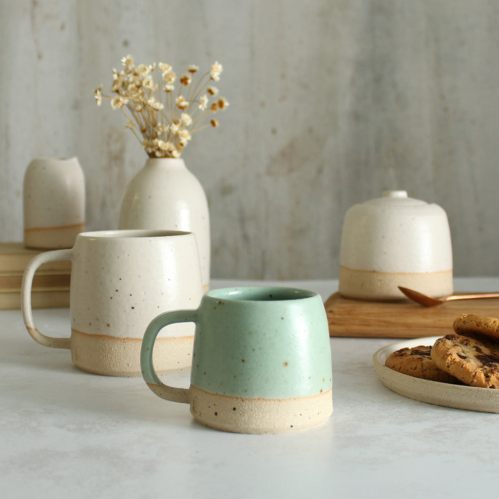 Habulous small stoneware mug mint green in front of large white mug and table setting of white stoneware ceramics