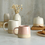 Habulous small stoneware mug pale pink in front of large white mug and table setting of white stoneware ceramics