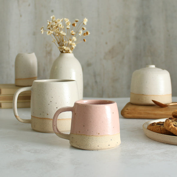 Habulous small stoneware mug pale pink in front of large white mug and table setting of white stoneware ceramics