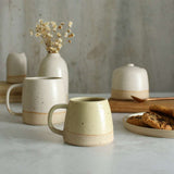 Habulous small stoneware mug light yellow in front of large white mug and table setting of white stoneware ceramics