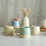 Collection of 5 pastel coloured small mugs on table with vase of flowers in background