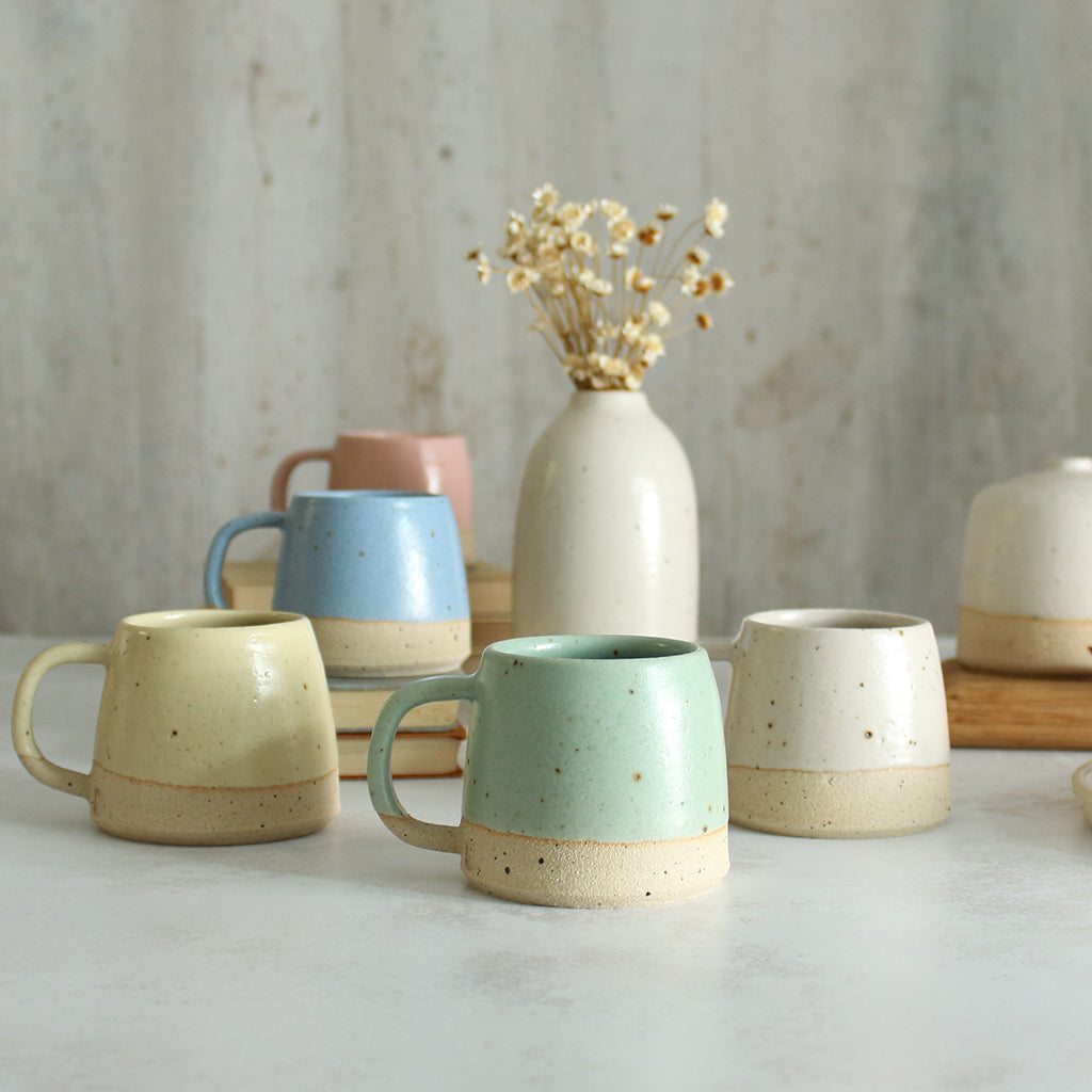 Collection of 5 pastel coloured small mugs on table with vase of flowers in background
