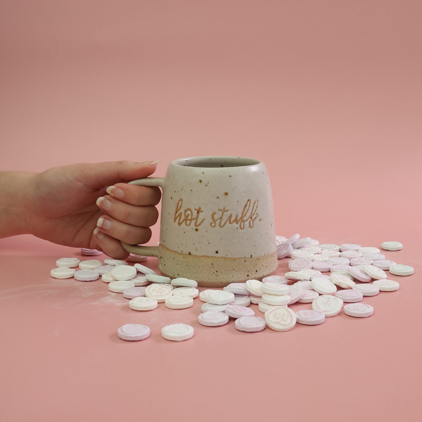 Hand holding a handmade stoneware mug in a white glaze labeled 'hot stuff' with candy love hearts surrounding the mug on a pink background