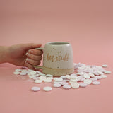 Hand holding a handmade stoneware mug in a white glaze labeled 'hot stuff' with candy love hearts surrounding the mug on a pink background