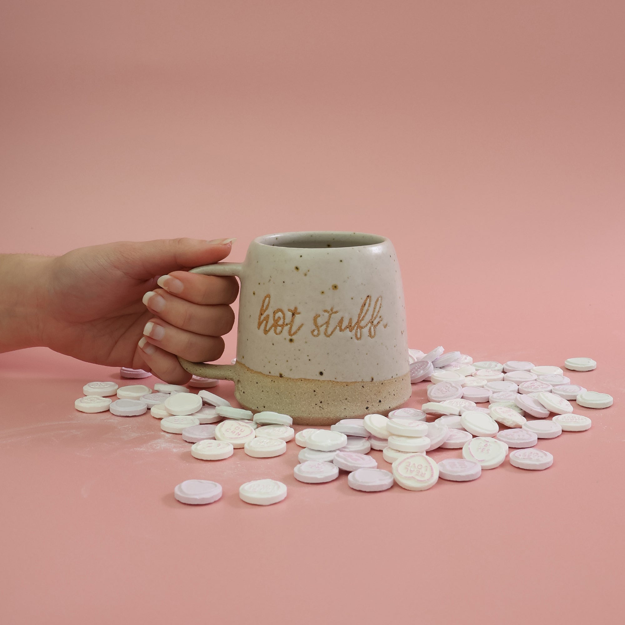 Hand holding a handmade stoneware mug in a white glaze labeled 'hot stuff' with candy love hearts surrounding the mug on a pink background