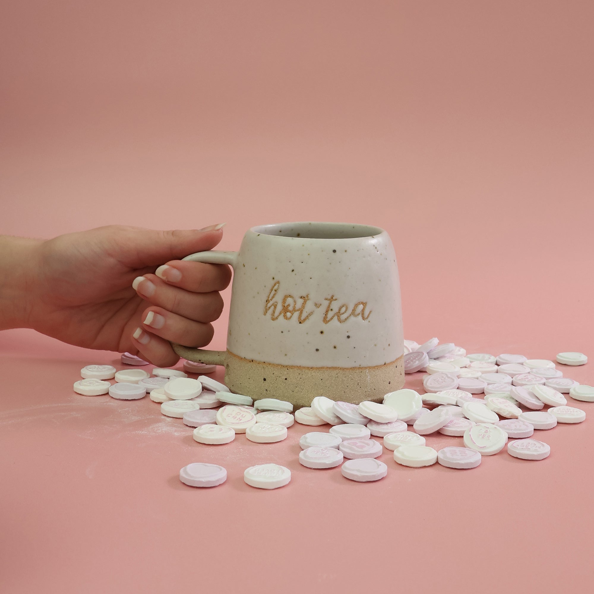 Hand holding a white handmade 'hot tea' mug with colorful candies on a pink background