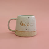 White ceramic mug handmade with fleck clay with 'hot tea' text on a pink background. a pun on the saying Hottie
