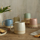 Our handmade ceramic mugs on a wooden table with a neutral background. The mugs are shown in five colours: yellow, cornflower blue, off-white, mint green and pale pink. 