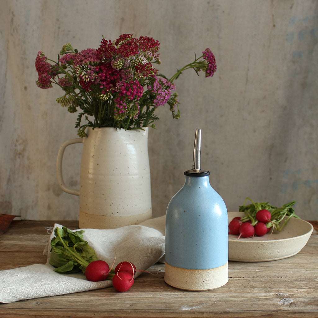 Small blue oil pourer on table next to a bowl of radishes and a jug of wildflowers